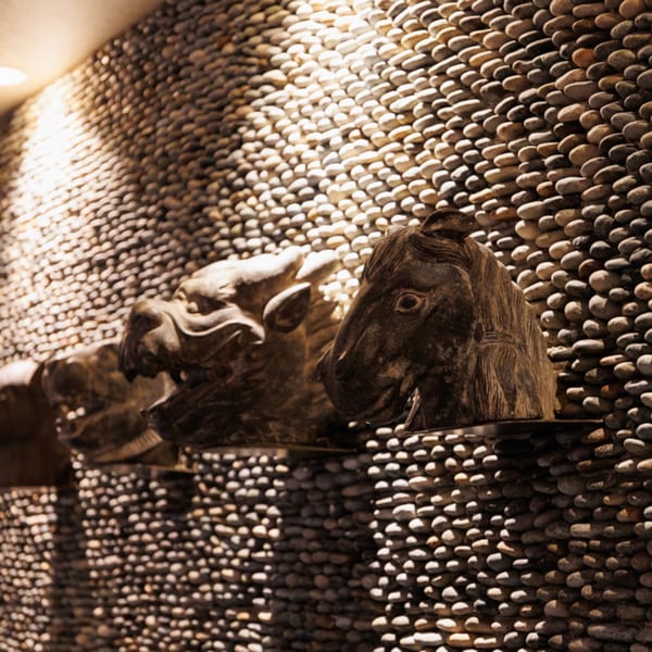 Carved wooden animal heads adorn a textured pebble wall, illuminated by warm lighting at Aja Asian Cuisine.