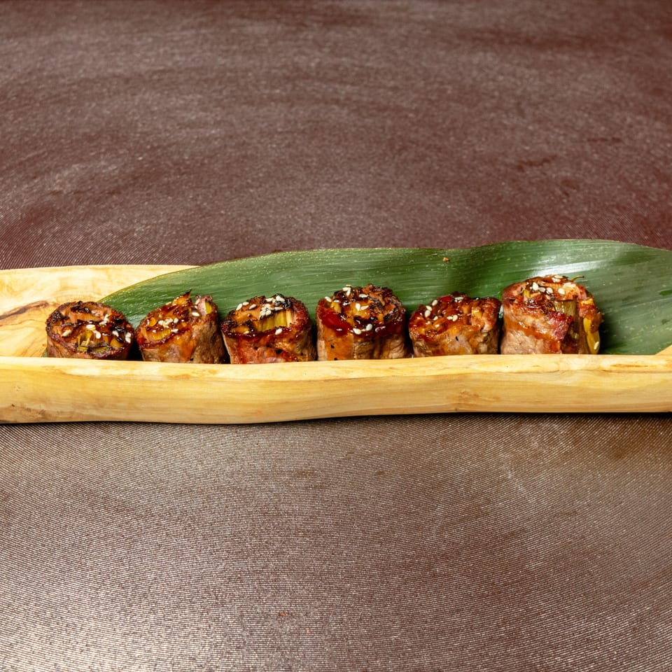 Six Asian beef rolls with sesame seeds and sauce, served on a wooden tray with a green leaf.