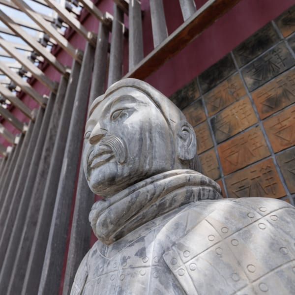 Stone statue of an Asian warrior with a mustache, against a red wall with patterned tiles.