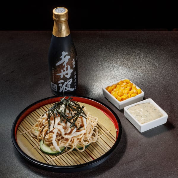 Asian noodles with seaweed and white strips, served with corn, sauce, and a sake bottle.
