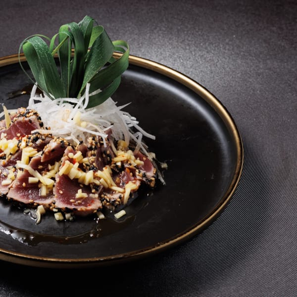 Seared tuna sashimi with sesame seeds, diced ginger, shredded daikon, and green garnish on a black plate.